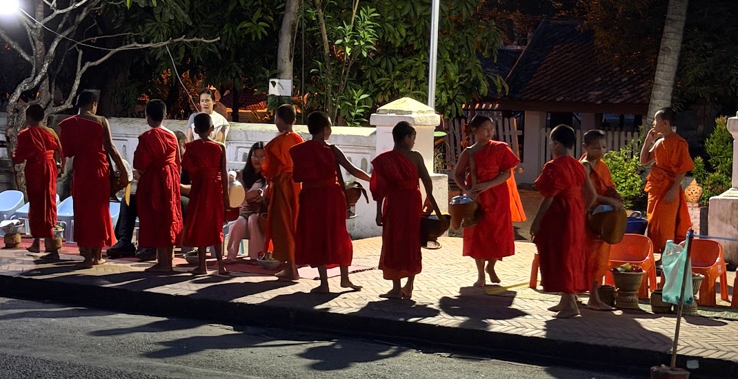 Tak Bat in Luang Prabang: eine Reihe barfüßiger Mönche in orangen Roben sammelt im Morgengrauen Almosen auf der Straße