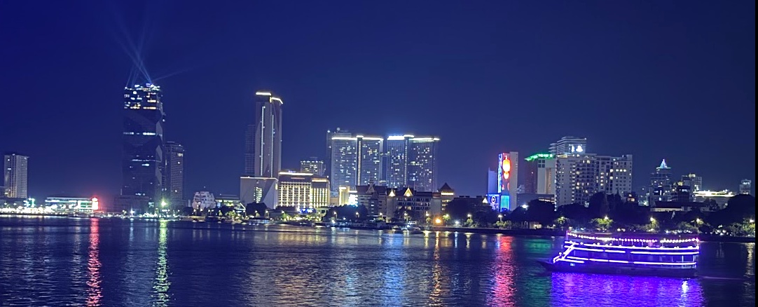 Skyline von Phnom Penh bei Nacht, Blick vom Fluss auf beleuchtete Hochhäuser und ein vorbeifahrendes Ausflugsboot